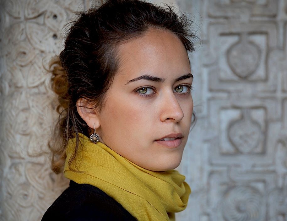 Portrait of an individual with dark hair pulled back, wearing a black top and mustard yellow scarf, looking intently at the camera against a textured stone background.