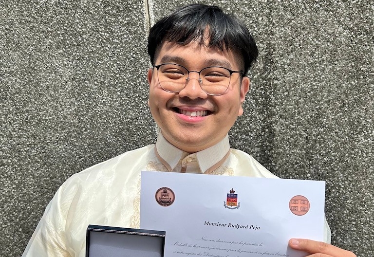 A young man with dark hair and glasses, smiling, holding a certificate