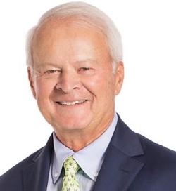 A man with short white hair smiles and is wearing a navy blue blazer over a light blue dress shirt and light green tie