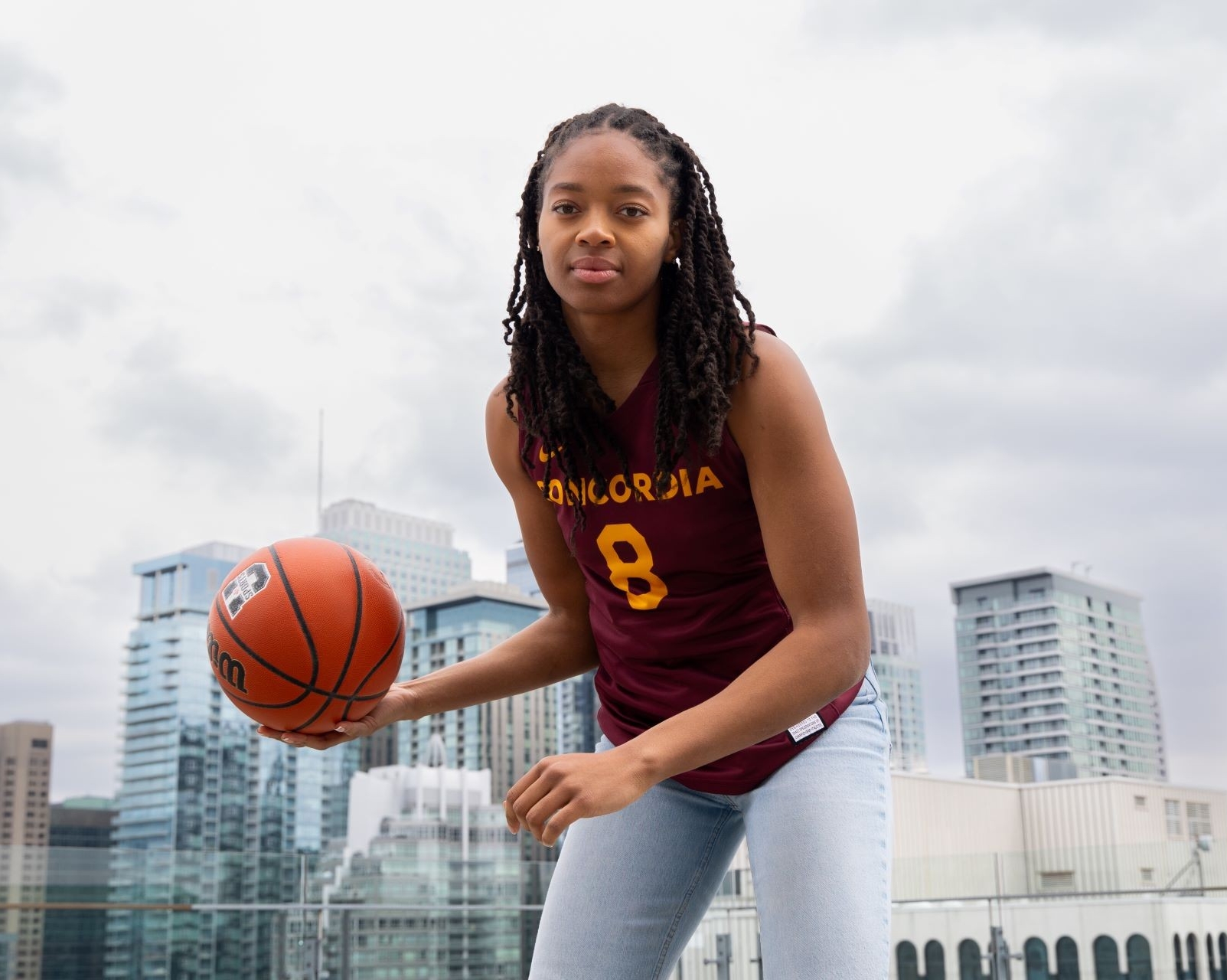 A female basketball player in Concordia uniform posing with a basketball, with Montreal's skyline in the background.