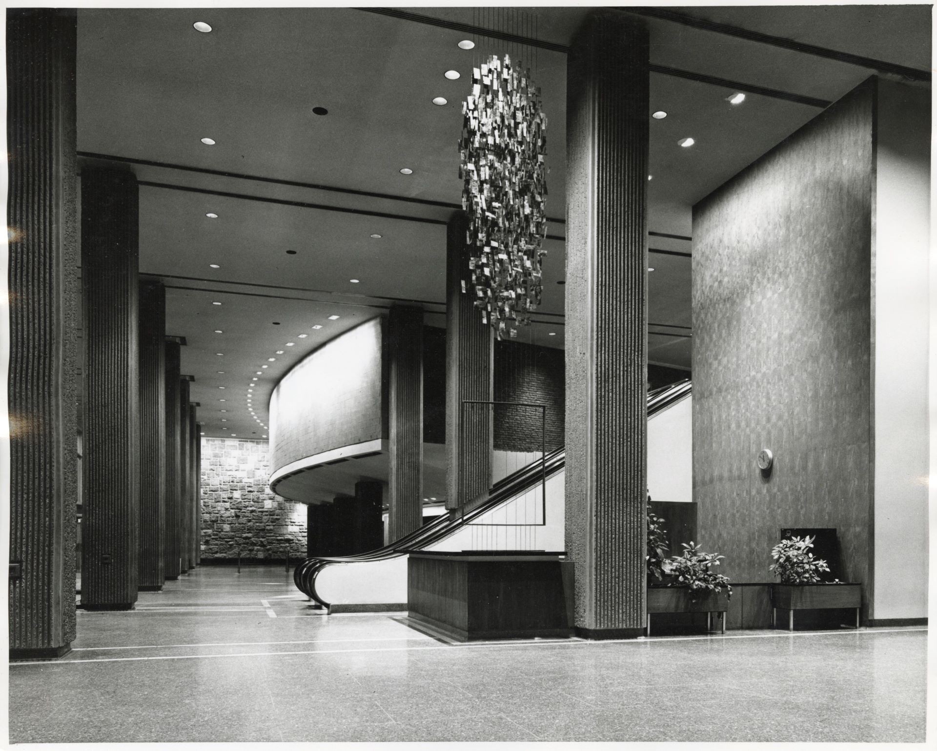 A vintage black and white photo of an elegant interior hall with a curving staircase and decorative light fixtures, reflecting mid-century modern design.