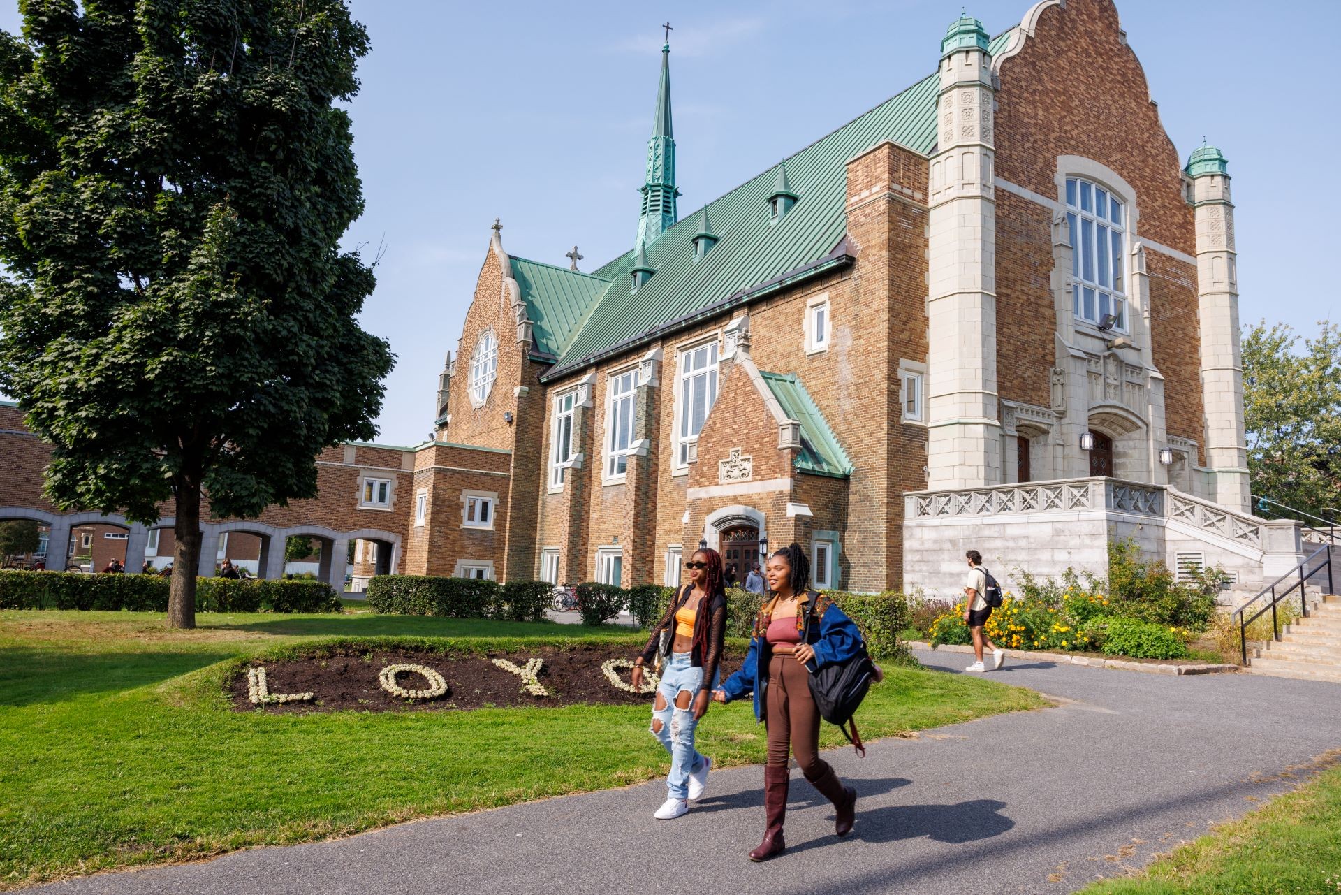 Two students walking past the historic Loyola campus building with "LOY" spelled out in a flower bed in the foreground.