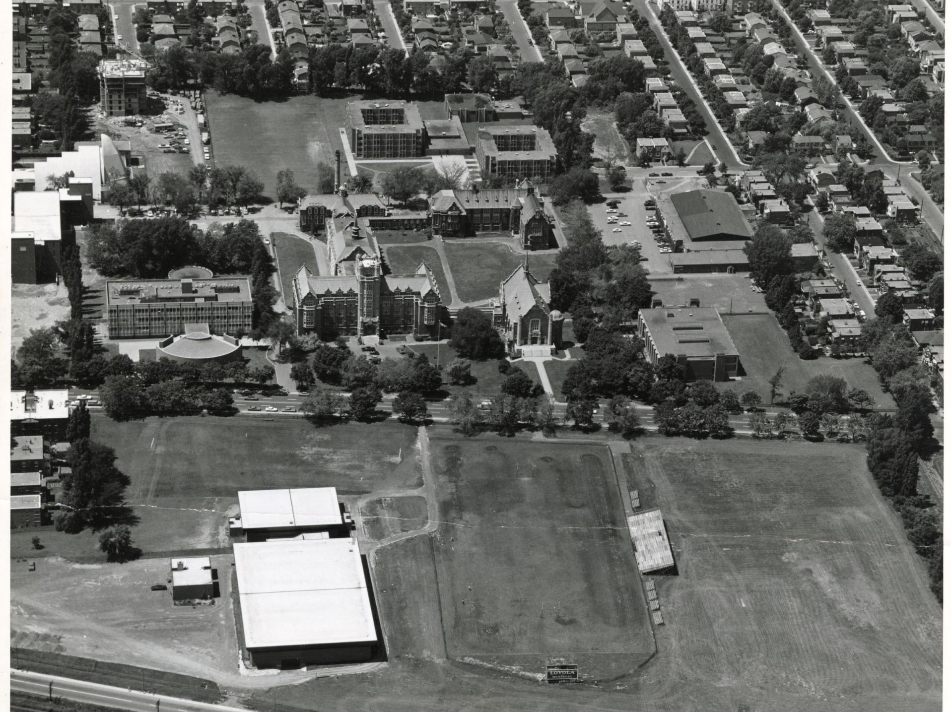 An aerial black and white photo of a campus with several buildings and surrounding residential streets, showing an extensive open area.