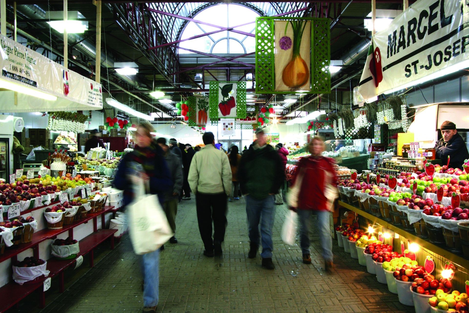 A bustling indoor market scene with vendors and customers around stalls filled with fresh produce and other goods.