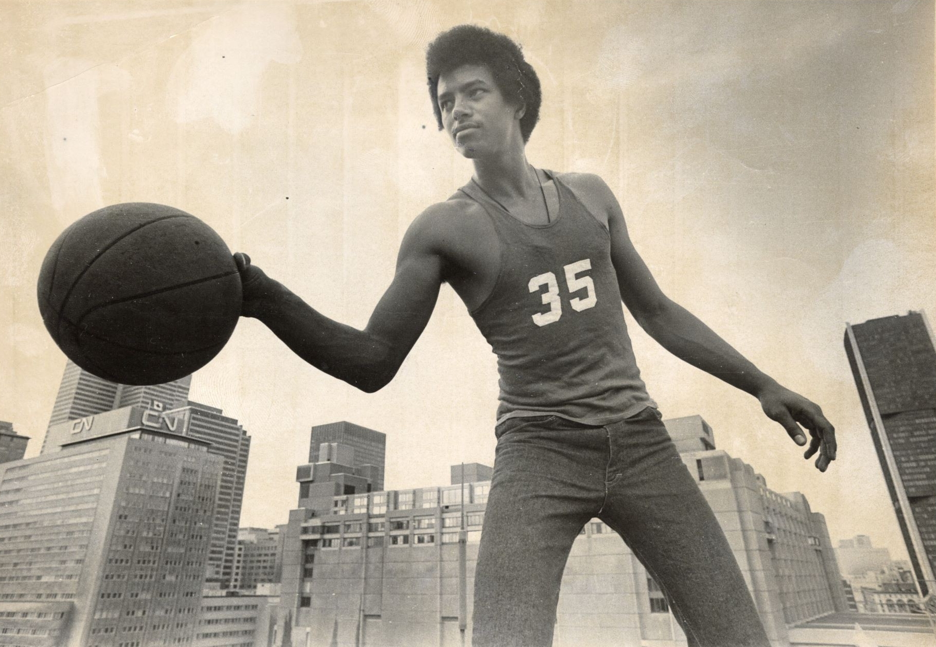 A vintage black and white photo of a young man holding a basketball with city skyscrapers in the background, evoking a sense of historical sports achievement.