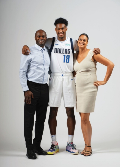 Two adults flank a young athlete wearing a "DALLAS 18" basketball uniform, all smiling proudly for the camera in a studio setting.