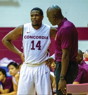 An individual in a white basketball uniform labeled "CONCORDIA 14" listens intently to a coach during a discussion on a basketball court with bleachers in the background.