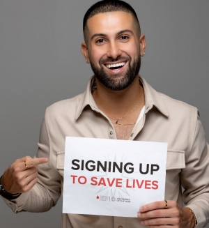 A person with short hair and a beard is smiling and holding a sign that reads: "Signing up to save lives"