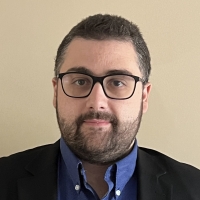 Image of a man with dark hair and beard wears a blue button up shirt under a black blazer. He is also wearing dark-frame glasses