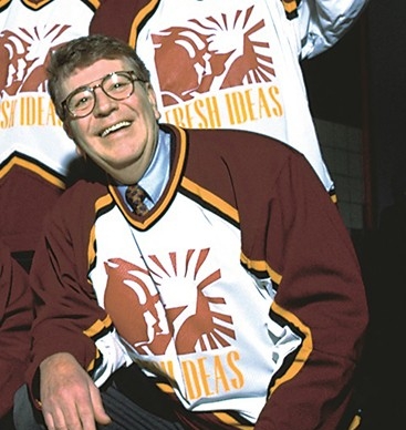 Image of a smiling man with brown, short hair, is wearing glasses and a hockey jersey with black dress pants