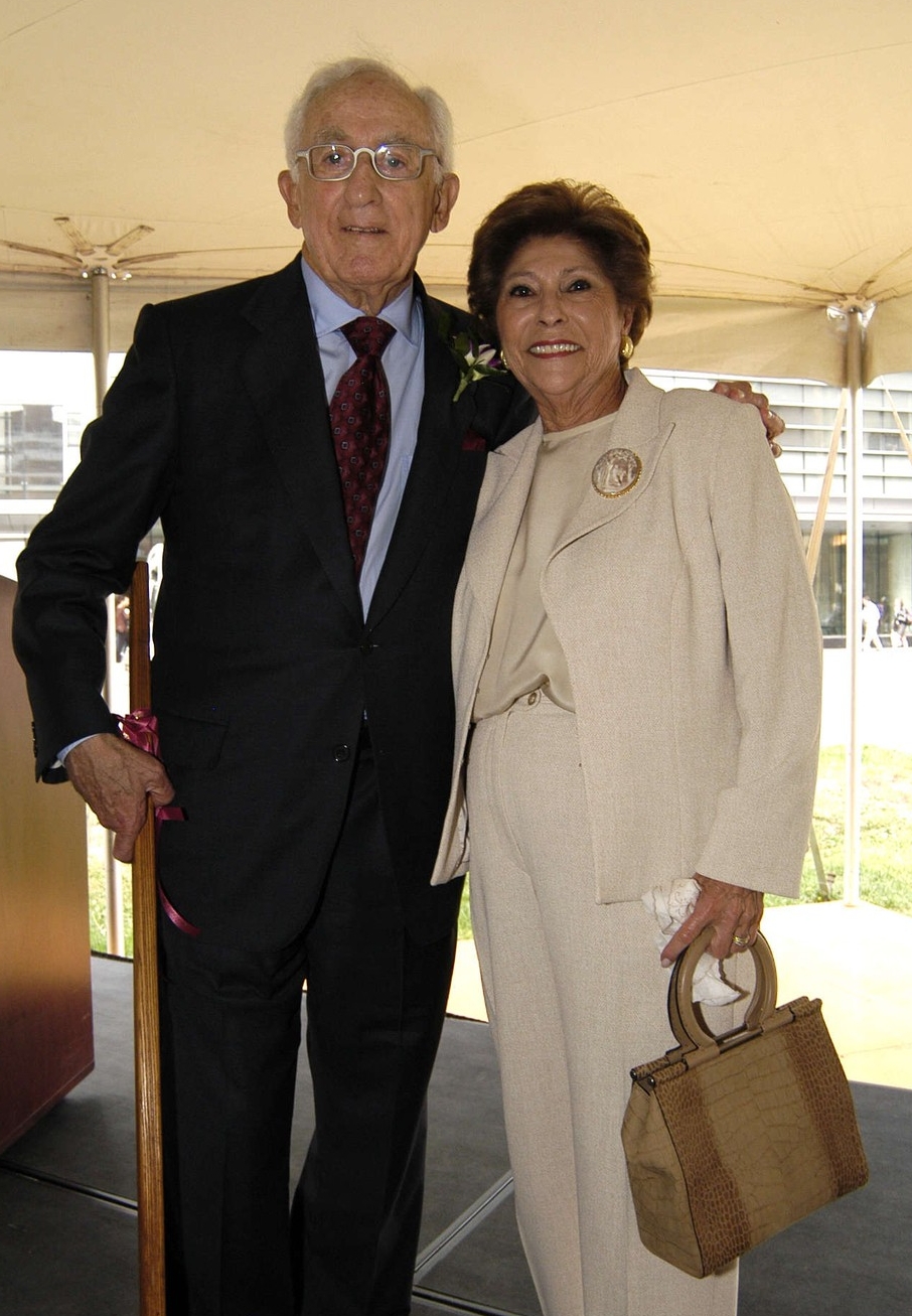 Photo of two people standing. A man wearing a dark suit and tie next to a woman wearing a white suit and holding a brown handbag. 