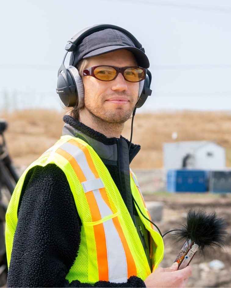 A student looks at the camera, wearing a safety vest and headphones while holding a sound recorder