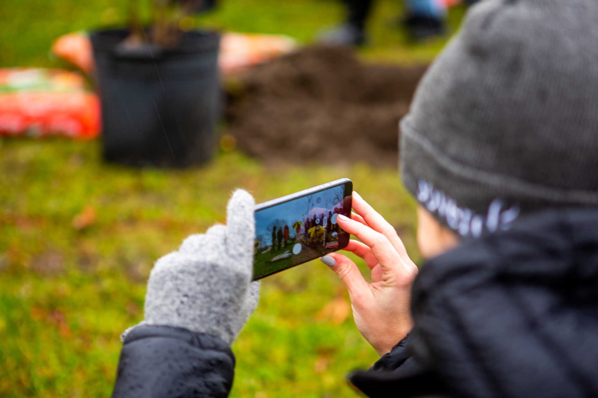 Une personne portant des gants prend une photo d'un événement de plantation d'arbres à l'aide d'un smartphone.