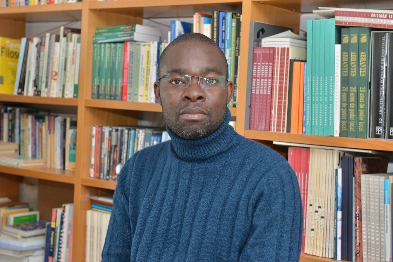 Visiting scholar Domingos da Cruz pictured in front of bookshelves wearing blue turtleneck and wire-rimmed glasses.