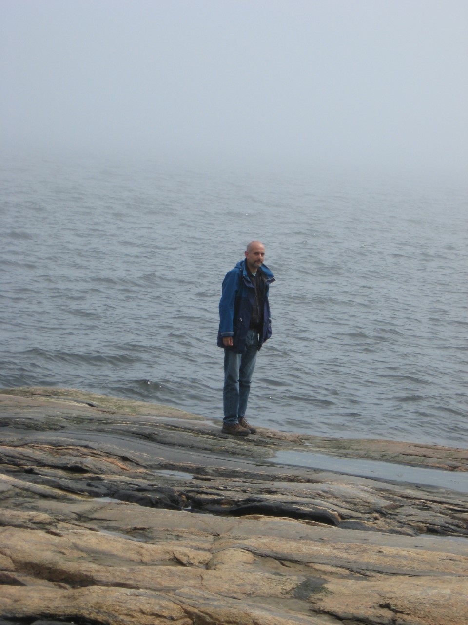 Aziz Choudry stands on a rocky beach with a large body of water behind him. He wears a dark blue rain coat and blue jeans.