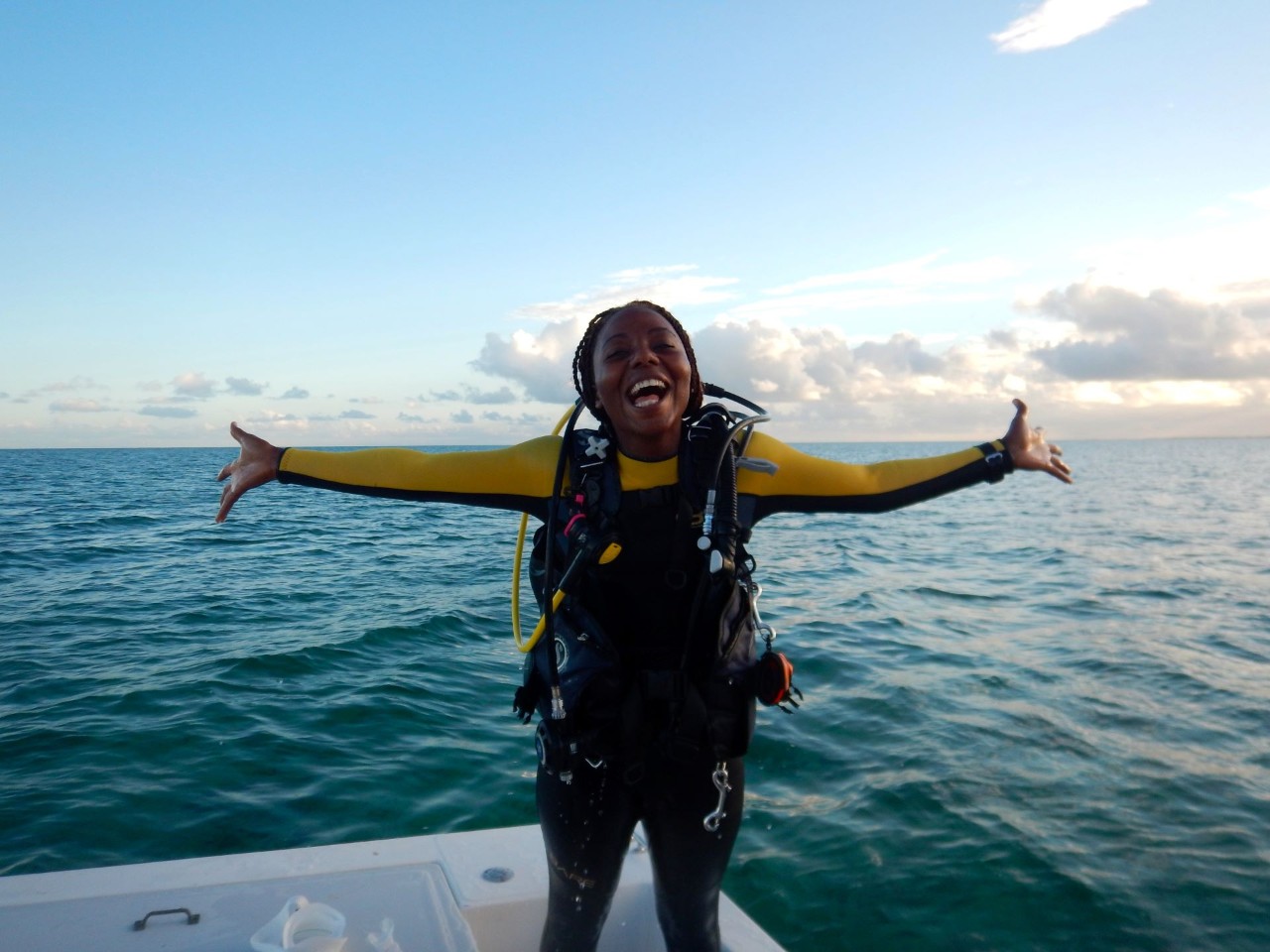 A Black woman smiling, standing on a boat with her arms spread out, wearing diving equipment