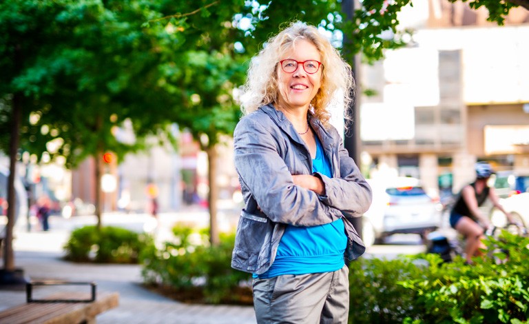 Woman with long, curly blonde hair, red-rimmed glasses, standing on a city street in summer.