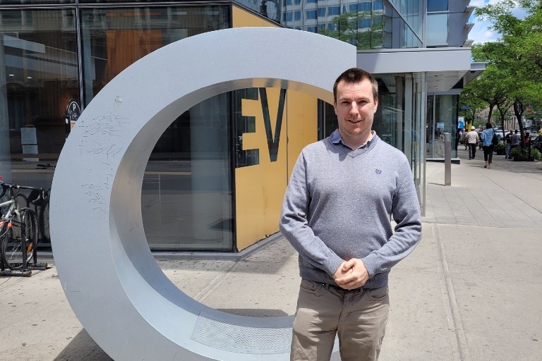 A man stands in front of a sculpture shaped like the letter c outside the EV Building on the Sir George Williams Campus.