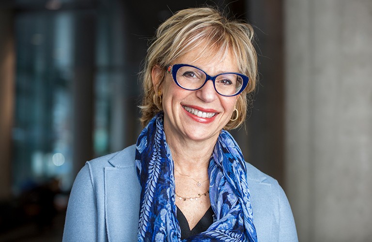 Pictured: Smiling woman with shortish blonde hair, glasses, a silk scarf and a blue shirt and jacket.