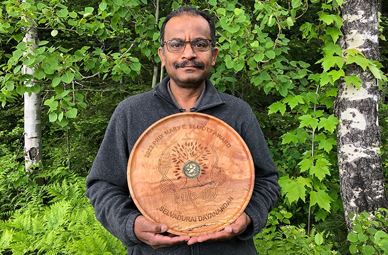 Older Indian gentleman standing in a forest and holding an award shaped like a round piece of wood.