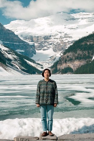 A woman in a sweater stands on a rock, with large snow-covered mountains in the background