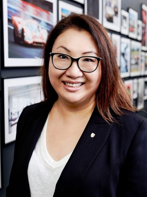 Kitty Luu has shoulder-length brown hair, wears black-framed glasses, and a navy blue blazer over a white shirt. She stands in front of a wall of framed photos of Porsche cars.