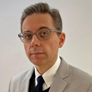 A man in a checkered suit wears glasses and looks at the camera