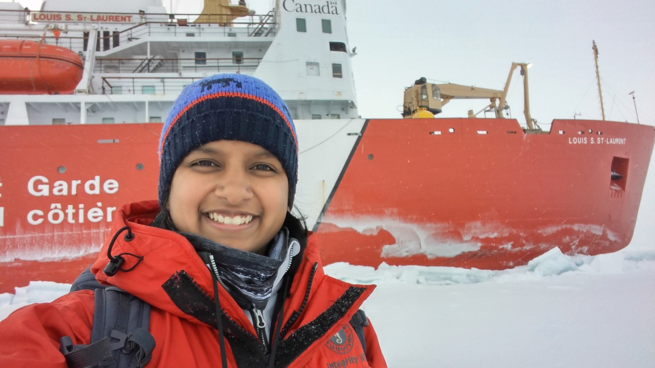 A woman wearing a colourful toque and red winter jacket stands smiling in front of a red icebreaker boat in the Arctic