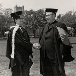 A young man shakes hands with an older man. They both wear graduation robes and square academic caps