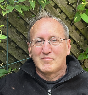 Headshot of a man with short grey hair and glasses leaning against a fence with green vines is wearing a black zip-up sweater
