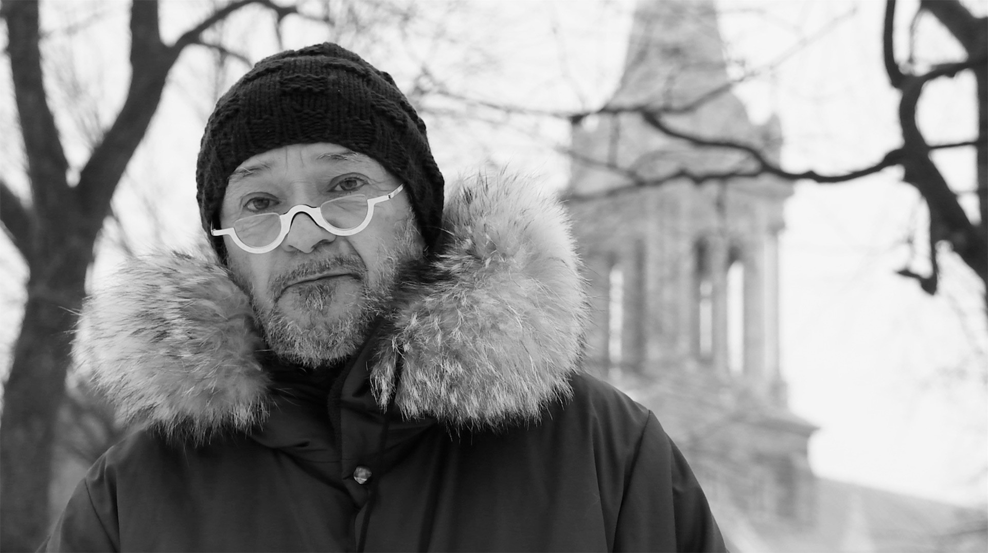 A black and whote image of a man wearing a winter parka, tuque and white-rimmed glasses