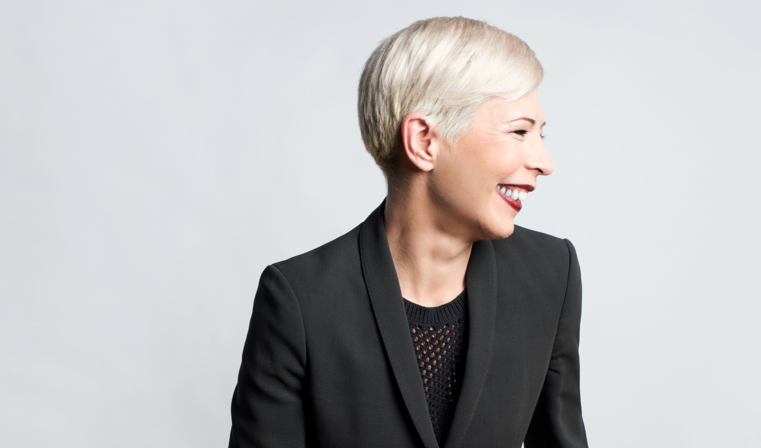 A woman with short, white-blonde hair stands in front of a white backdrop and looks off to the side while smiling. She wears burgundy lipstick, black nail polish, a silver ring and a black suit.