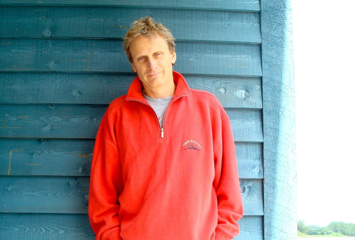 A man with light brown hair is wearing a bright red half-zip sweater over a grey t-shirt stands outside in front of teal-coloured wood siding
