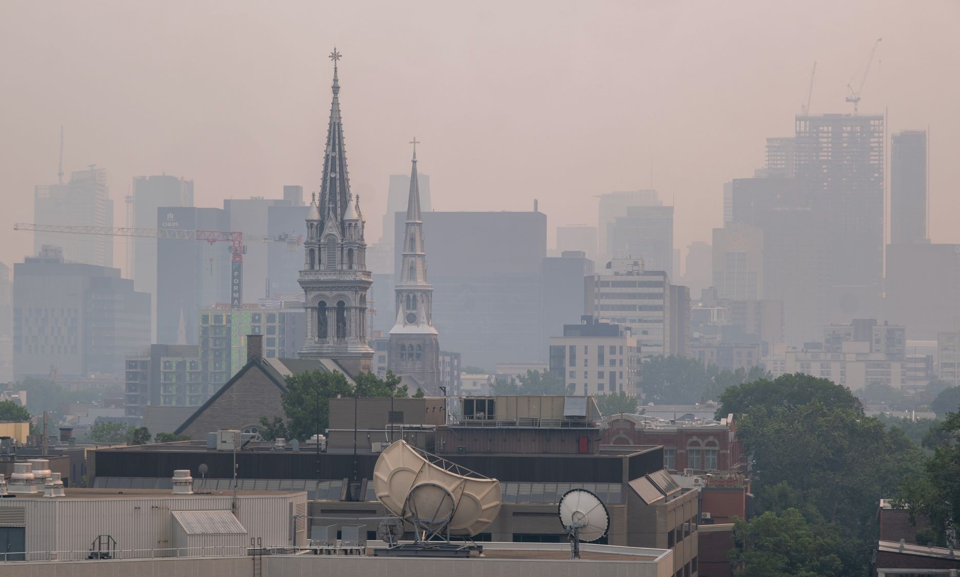 A cityscape obscured by a haze, with the spires of two churches prominently rising in the foreground. The background shows obscured buildings and construction cranes, indicating ongoing development. Satellite dishes and rooftop equipment are visible on the buildings in the foreground, contrasting the old and the new in an urban environment with air quality issues.