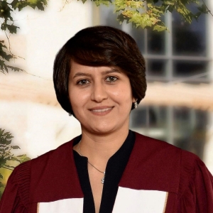 A woman with short hair is smiling and wearing academic regalia, which includes a maroon and white gown, typically worn for graduation ceremonies or other formal academic events. She is positioned outdoors with trees and a building with white window frames in the background.