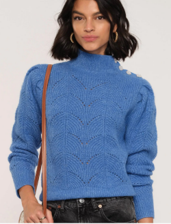 A woman with chin-length dark hair is wearing a sky-blue turtleneck sweater with black jeans. A long strapped beige purse hangs from her right shoulder