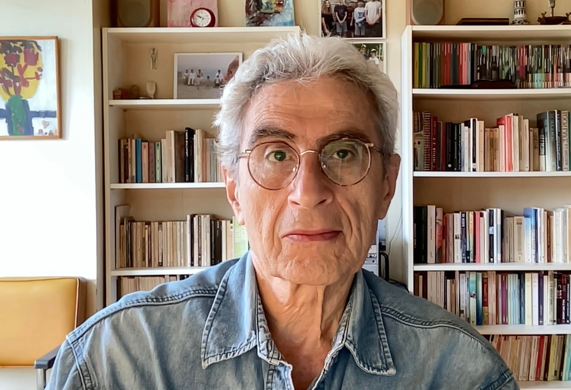 A man with white hair sits in front of bookshelves filled with books and framed photographs. He is wearing brown, round-framed glasses and a light jean shirt.