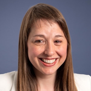 A woman with light brown hair smiles in front of a blue-grey background. She is wearing a cream-coloured blazer.