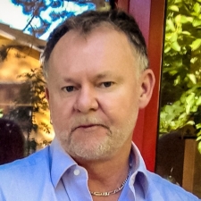 Portrait of a man with short hair and a beard, wearing a blue button-up shirt with a silver chain 