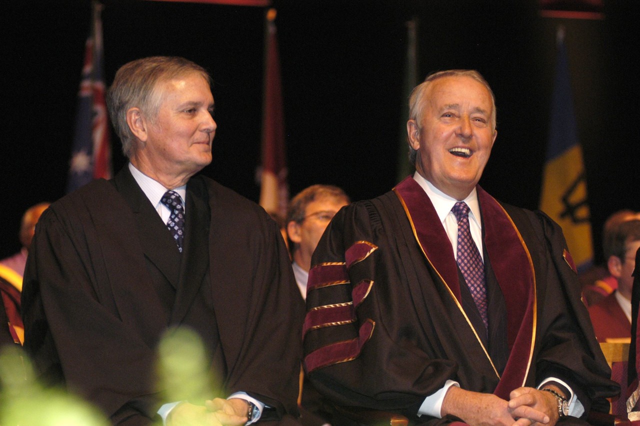 Two men sit smiling on stage dressed in graduation regalia