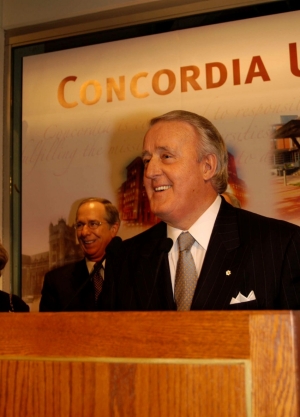 A man in a suit and tie stands at a podium to give a speech in front of a Concordia sign on the wall behind him.