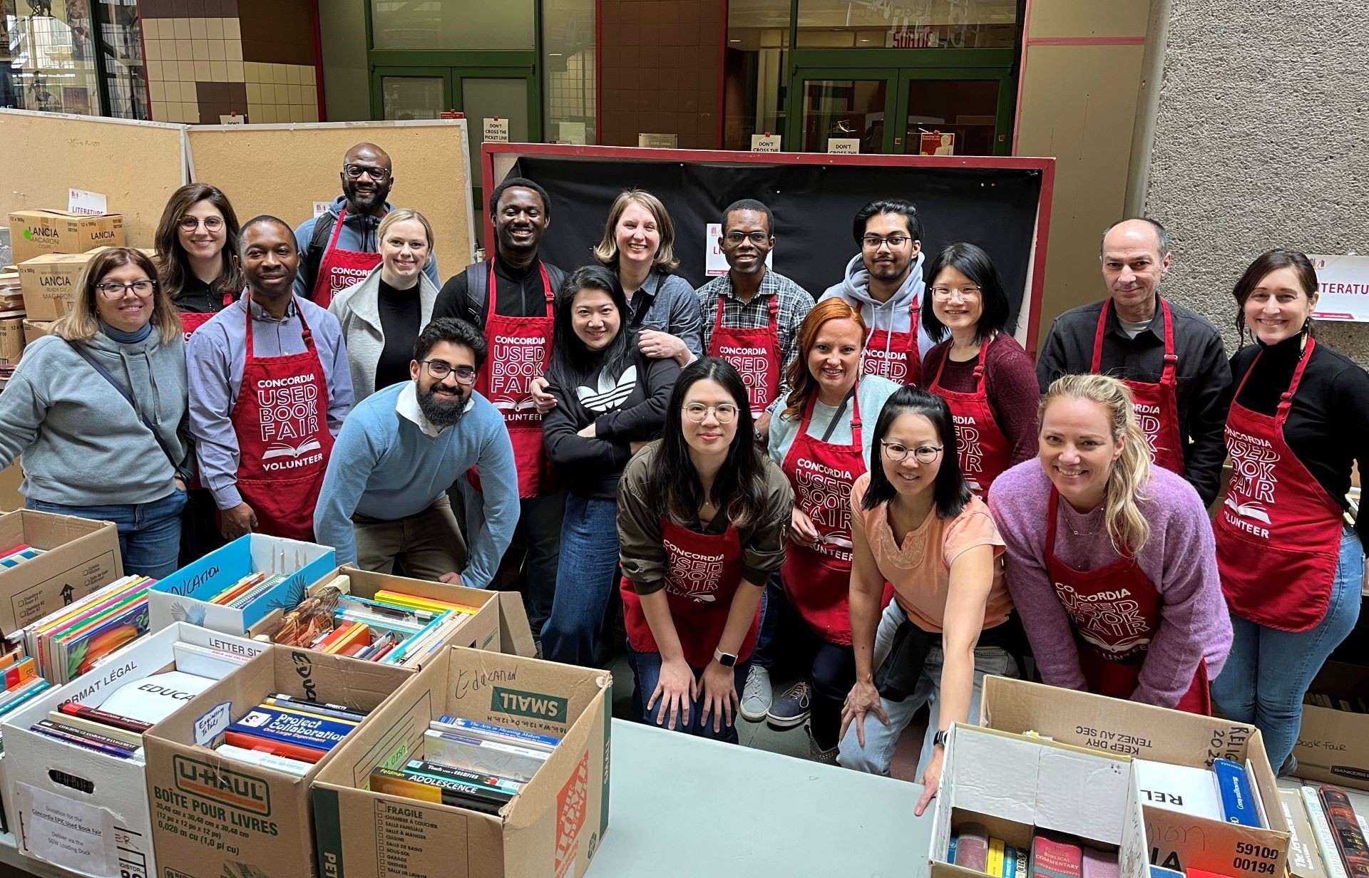 A group of cheerful volunteers wearing red aprons that read "Concordia Used Book Fair Volunteer" are standing behind boxes filled with books at an indoor event.