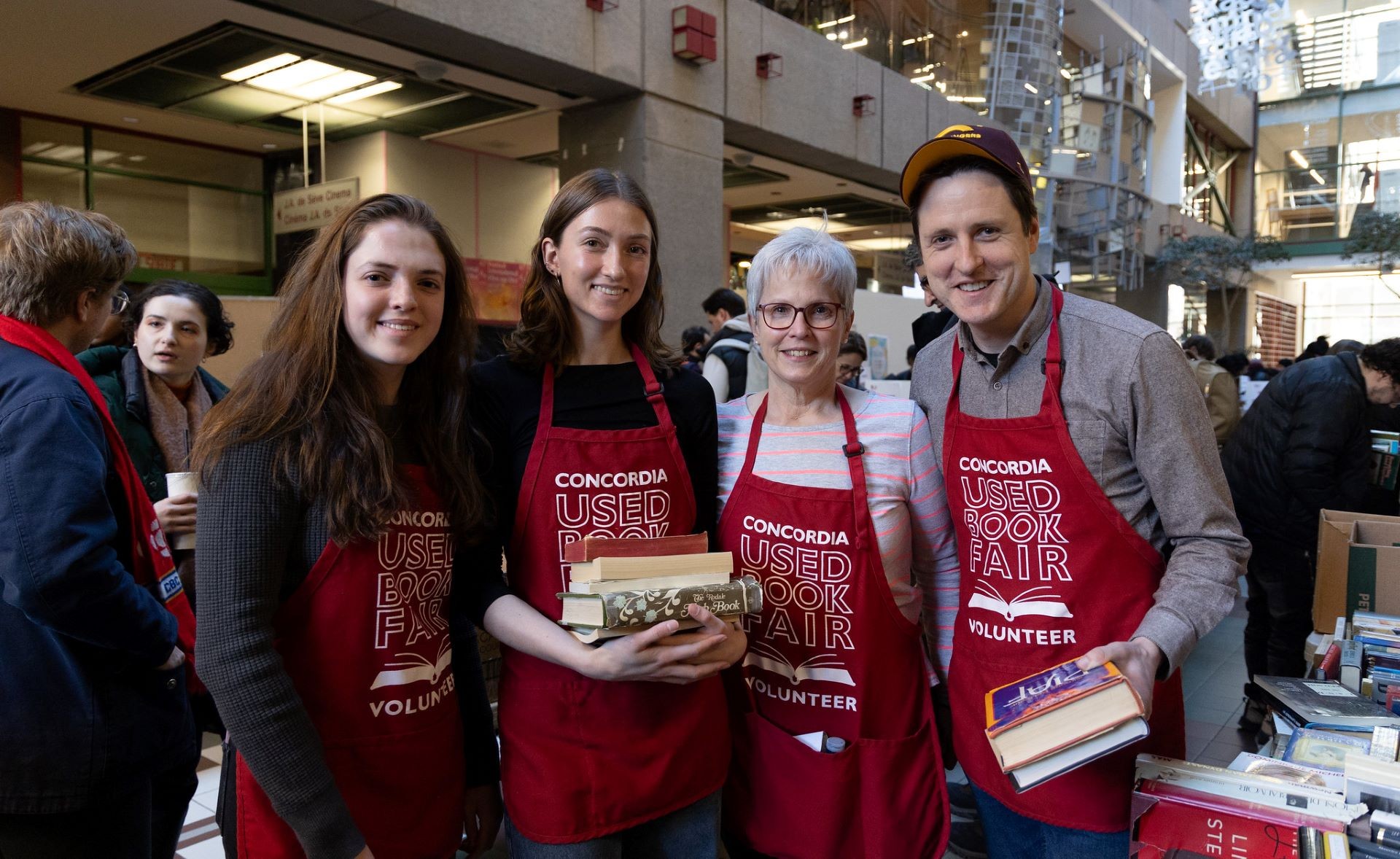 Four volunteers wearing red aprons with the text "Concordia Used Book Fair Volunteer" are smiling for the camera, holding a stack of books each, at an indoor book fair with other attendees in the background.