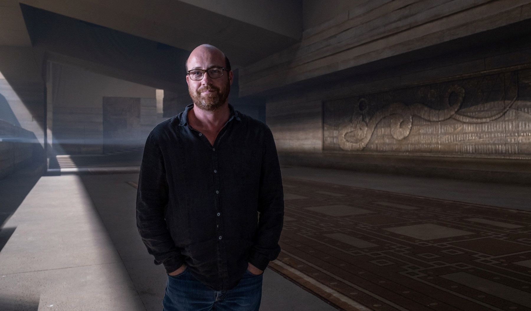 In a spacious, dimly-lit interior, a man in a black shirt stands confidently with hands in pockets, with striking beams of light and intricate wall engravings forming a dramatic backdrop.
