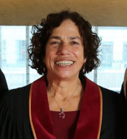 A woman with short, curly hair smiles at the camera. She is wearing a graduation gown