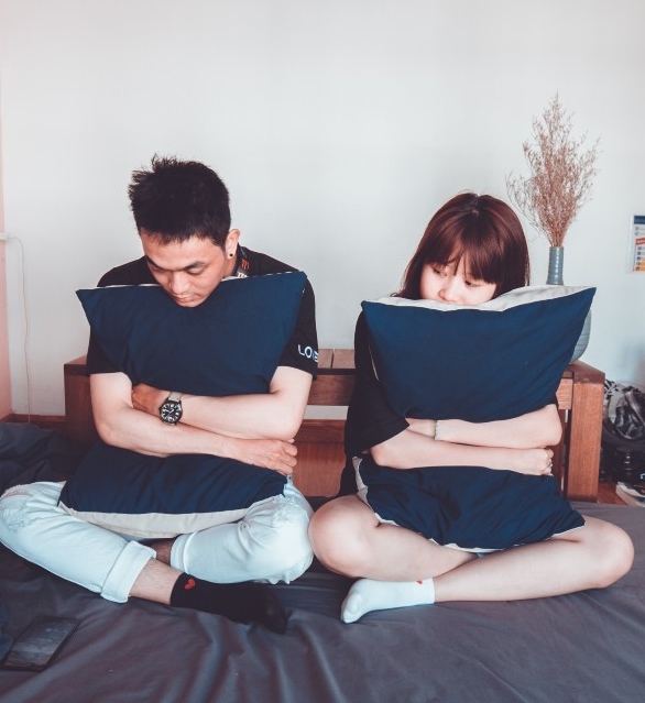 A young couple sits apart on a bed, each hugging a pillow