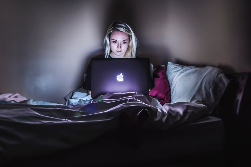A woman in a dark bedroom sits on her bed and uses a laptop