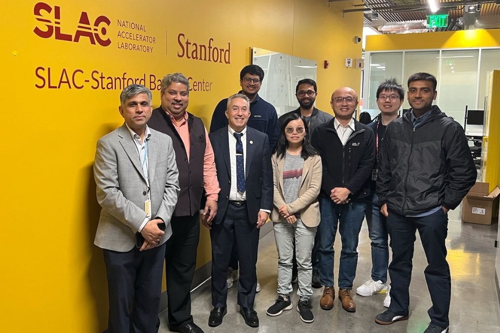 A group of scientists pose in front of a yellow wall with the words 'SLAC National Accelerator Laboratory Stanford' and below it the words 'SLAC-Stanford Battery Center'.