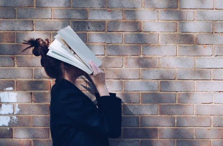 Une femme marchant par un mur de briques tenant un livre ouvert sur son visage