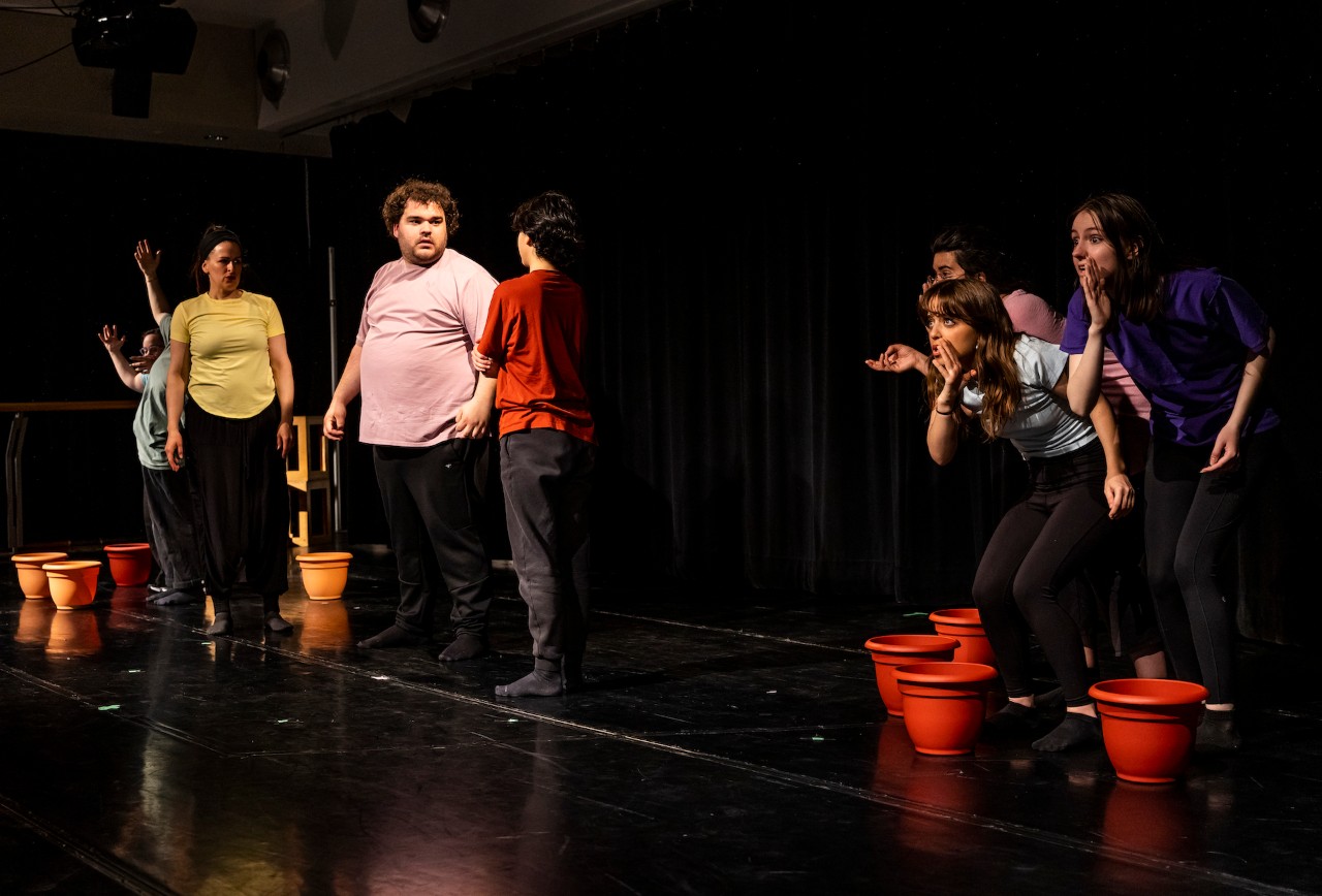 Plusieurs acteurs sur une scène de théâtre, avec des pots de fleurs éparpillés.